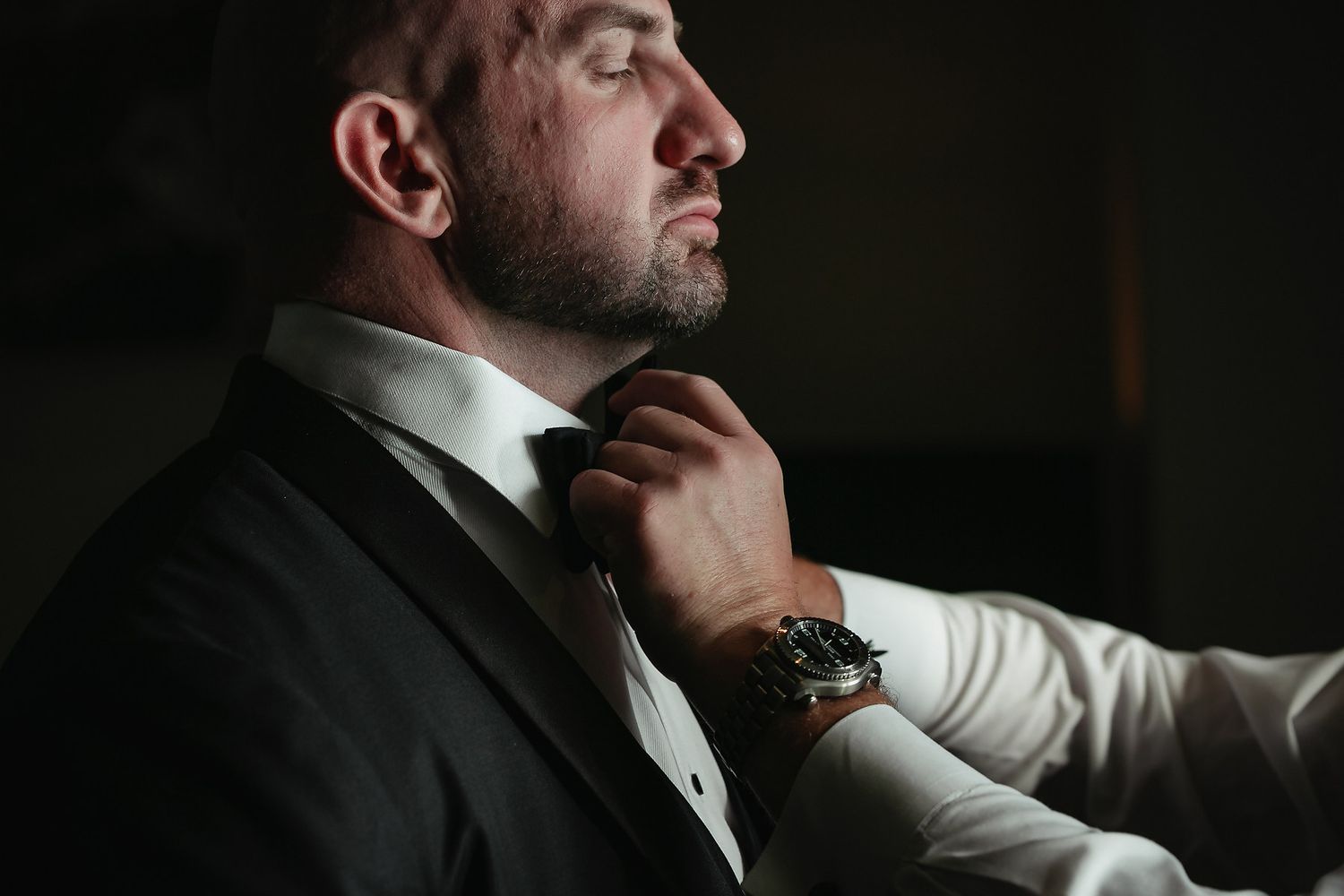 Profile view of a man in dark attire preparing for a formal event in moody lighting.