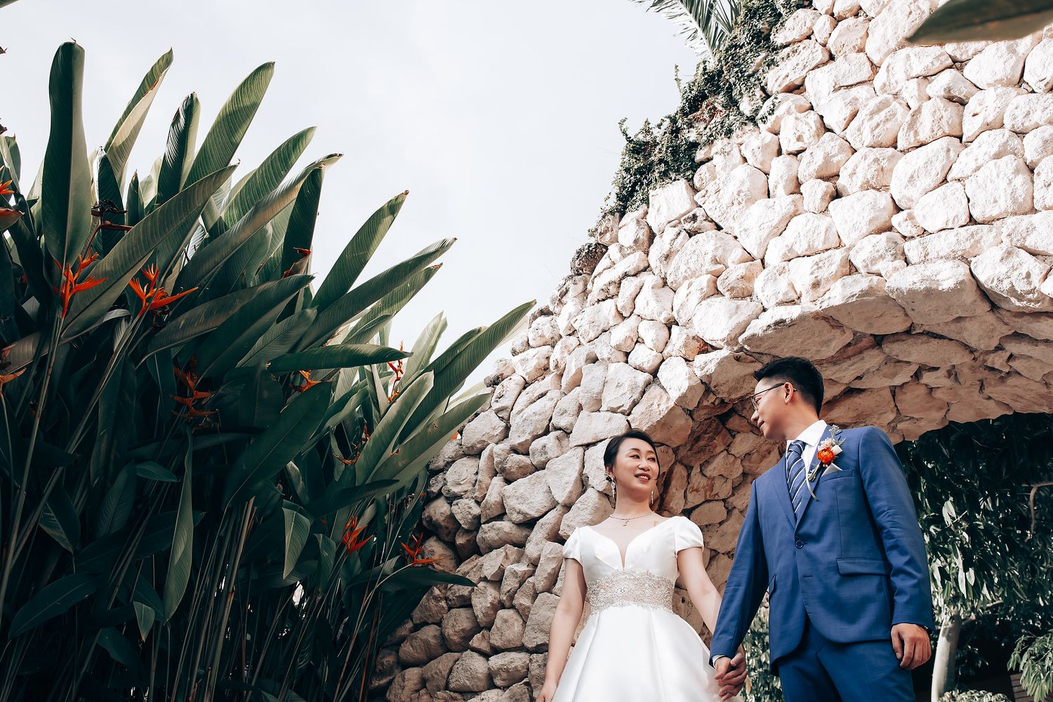 Wedding couple posing against a rustic stone wall with tropical plants.