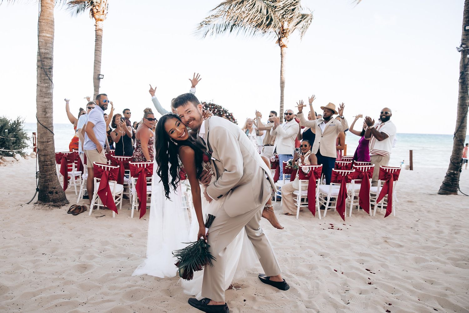 Newlyweds share a joyful dance moment on the beach while wedding guests cheer in the background.