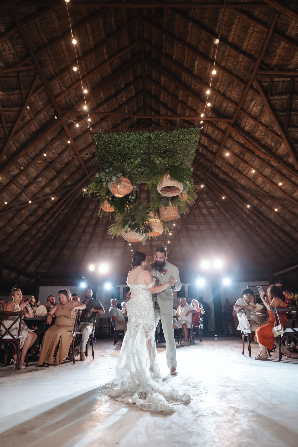 Bride and groom share first dance under string lights and tropical floral chandelier in a rustic thatched-roof wedding venue.