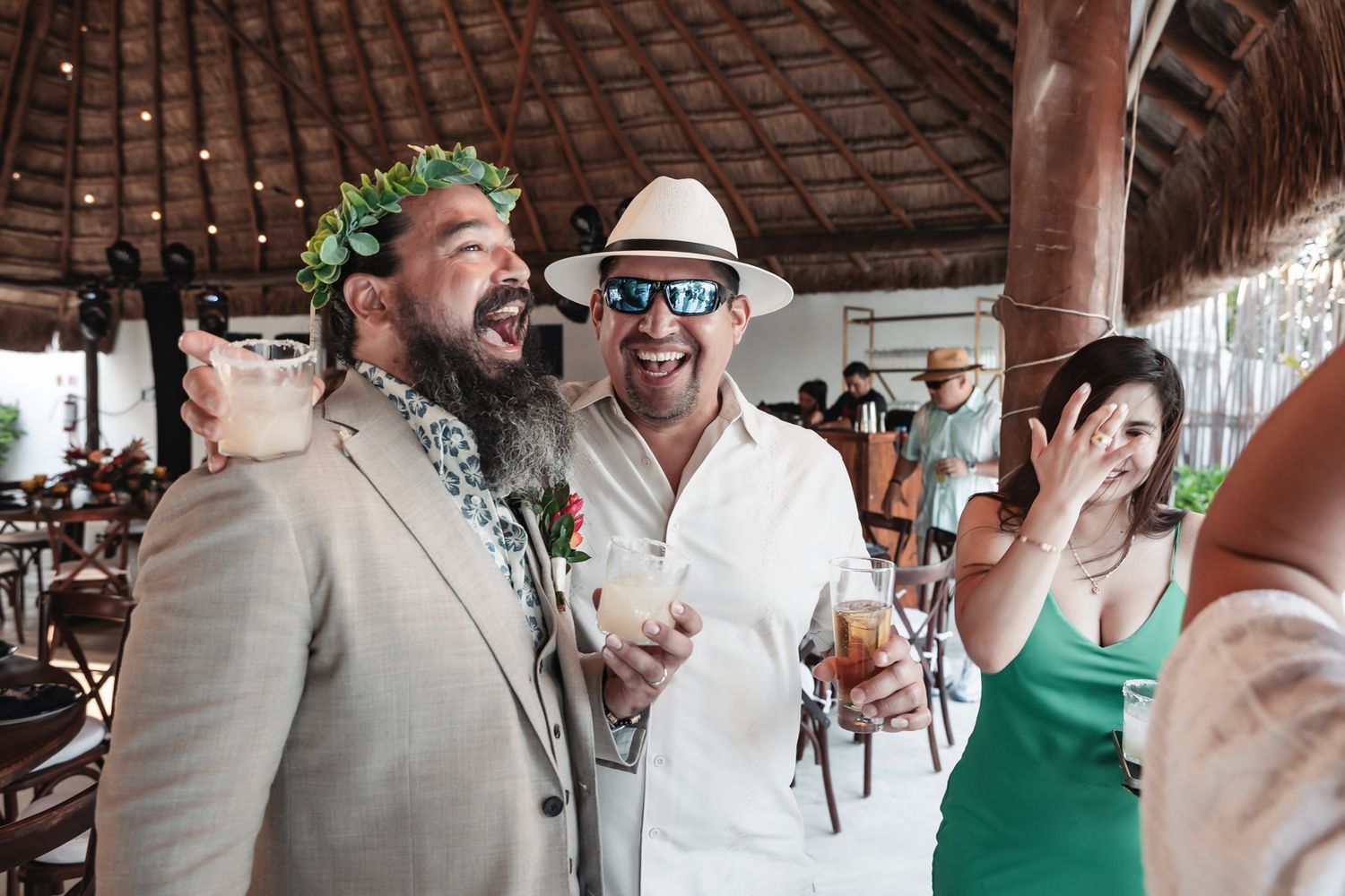 Two men laughing and celebrating at an outdoor tropical wedding reception, one wearing a floral crown and the other a white hat.