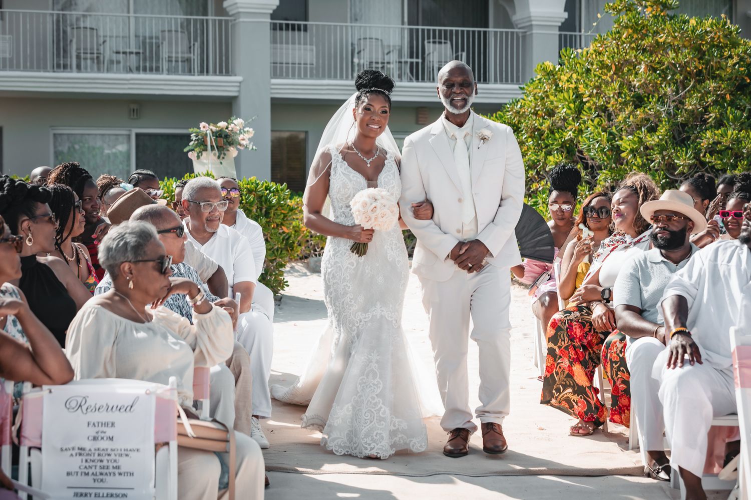 A joyful couple walks down the aisle at an outdoor beach wedding, bride in lace gown and groom in white suit.