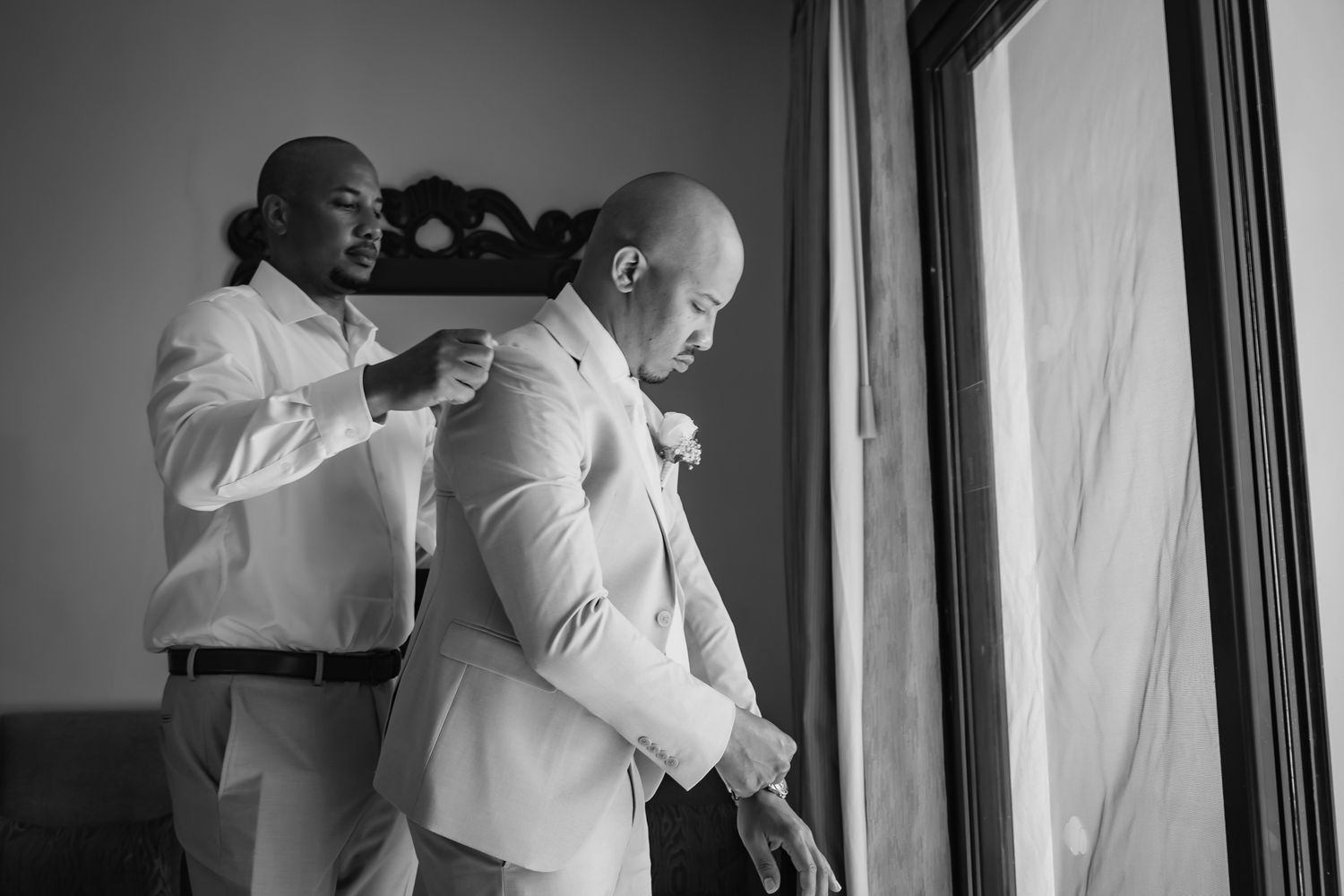 Groomsman helping bald groom with boutonniere near window in black and white wedding photo.