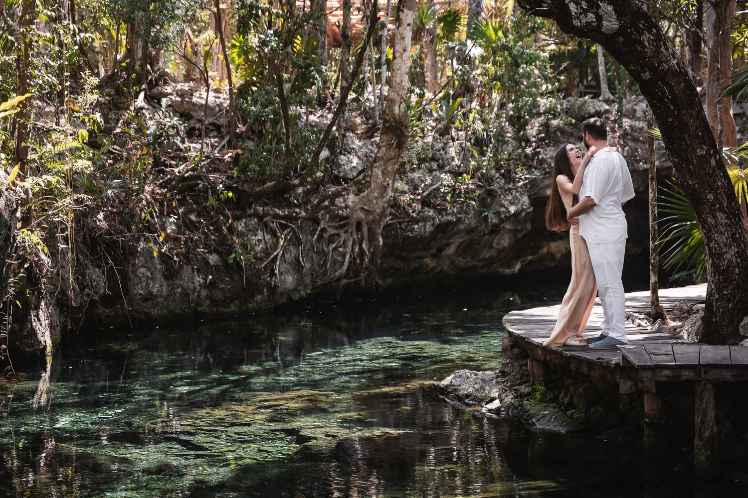 Romantic couple embracing on a wooden dock beside a serene cenote surrounded by lush tropical jungle.