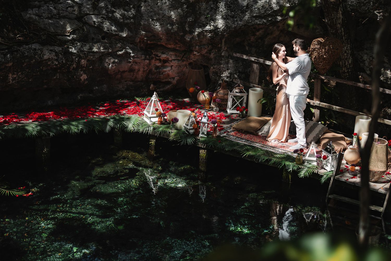Couple sharing a romantic moment on a decorated platform over a cenote surrounded by lush greenery and rose petals.