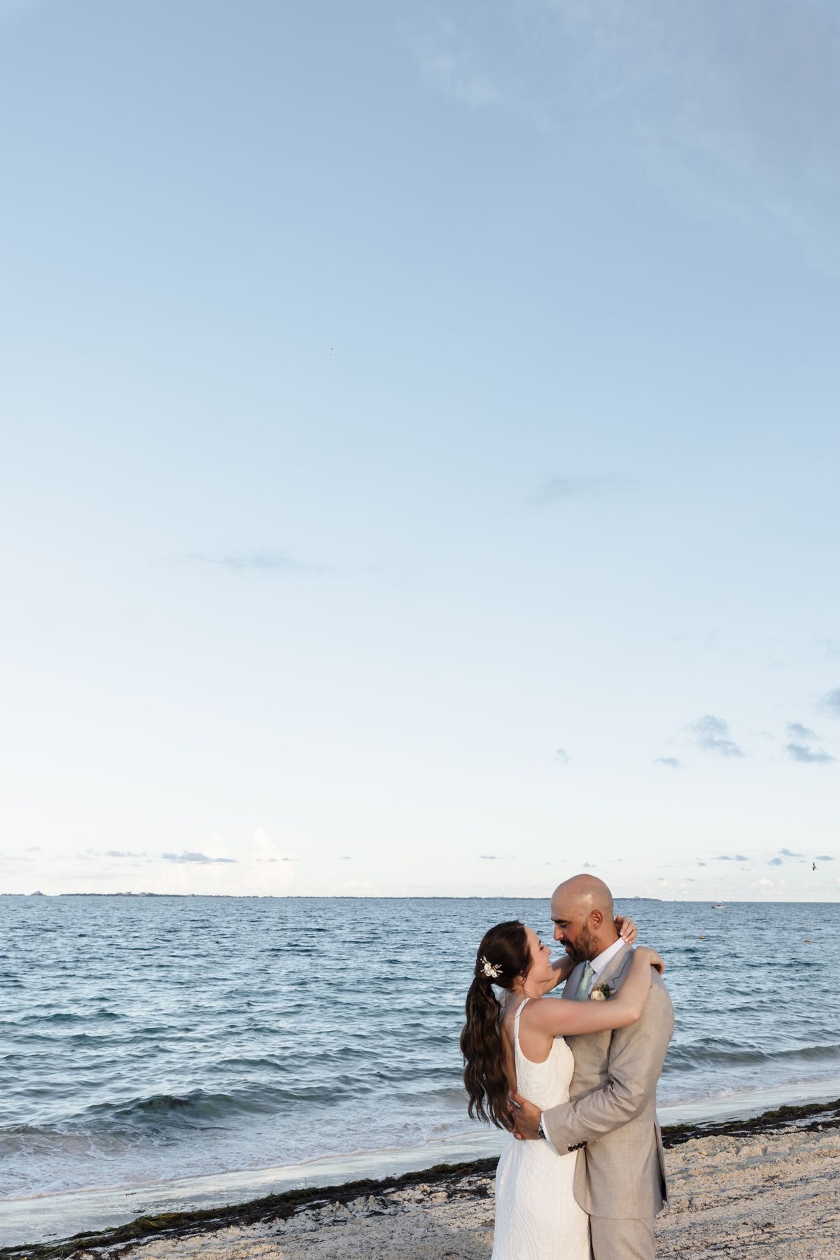 Bride and groom embrace on a beach at sunset, sharing a romantic moment by the ocean in wedding attire.
