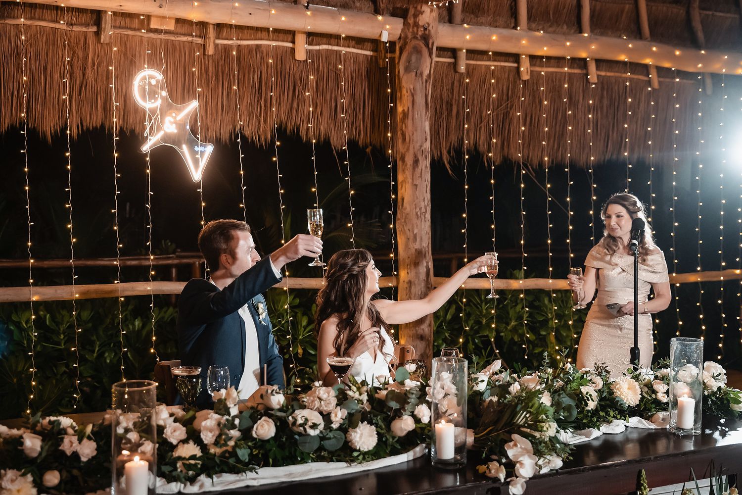 Wedding toast at a rustic barn reception with fairy lights, floral centerpieces, and candles as bride and guests raise glasses.