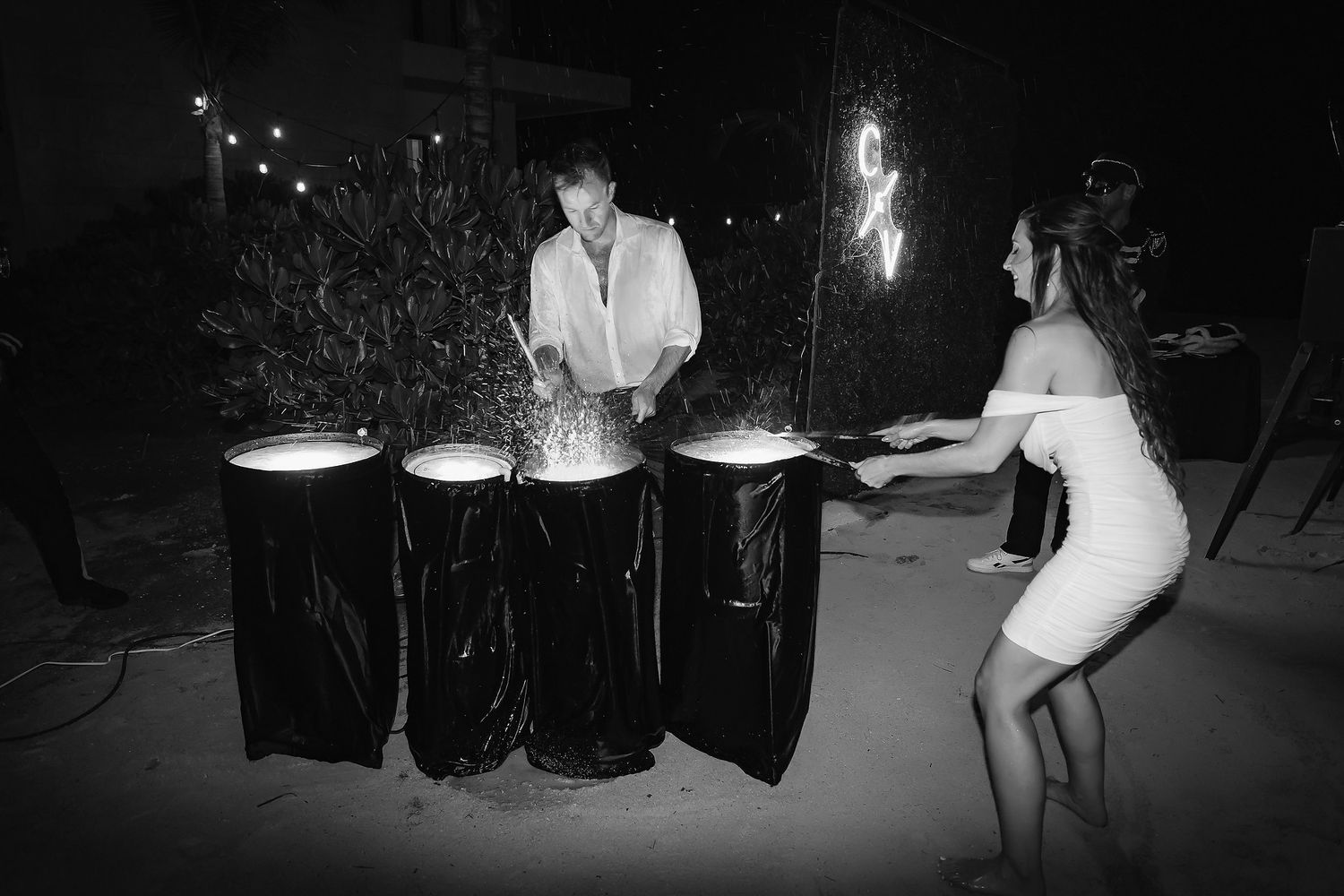 Black and white photo of two people lighting sparklers over draped tables at an outdoor night celebration.