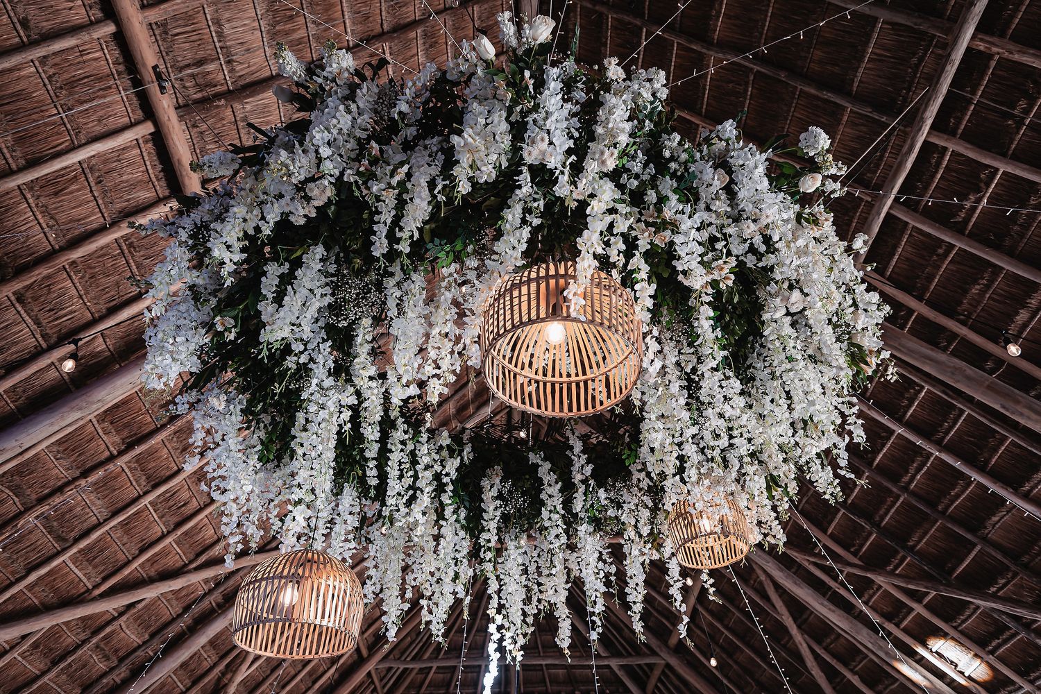 Elegant wedding ceiling decor with cascading white flowers and warm glowing rattan lanterns under a thatched roof.