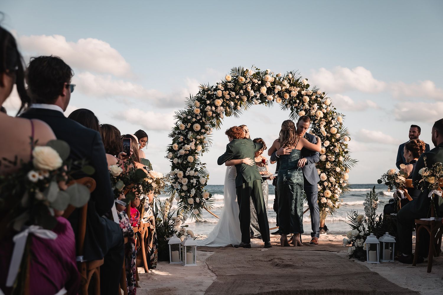 Couple embraces at outdoor beach wedding ceremony beneath a lush circular floral arch with guests seated alongside.