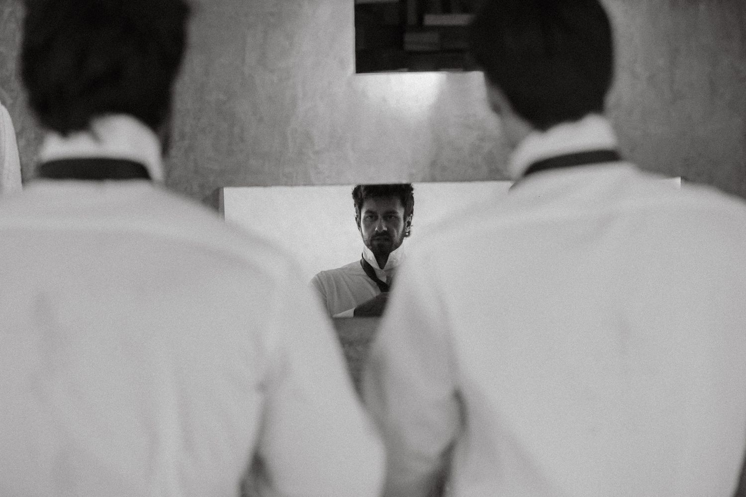 Black and white photo of a man reflected in a mirror between two people in white shirts.