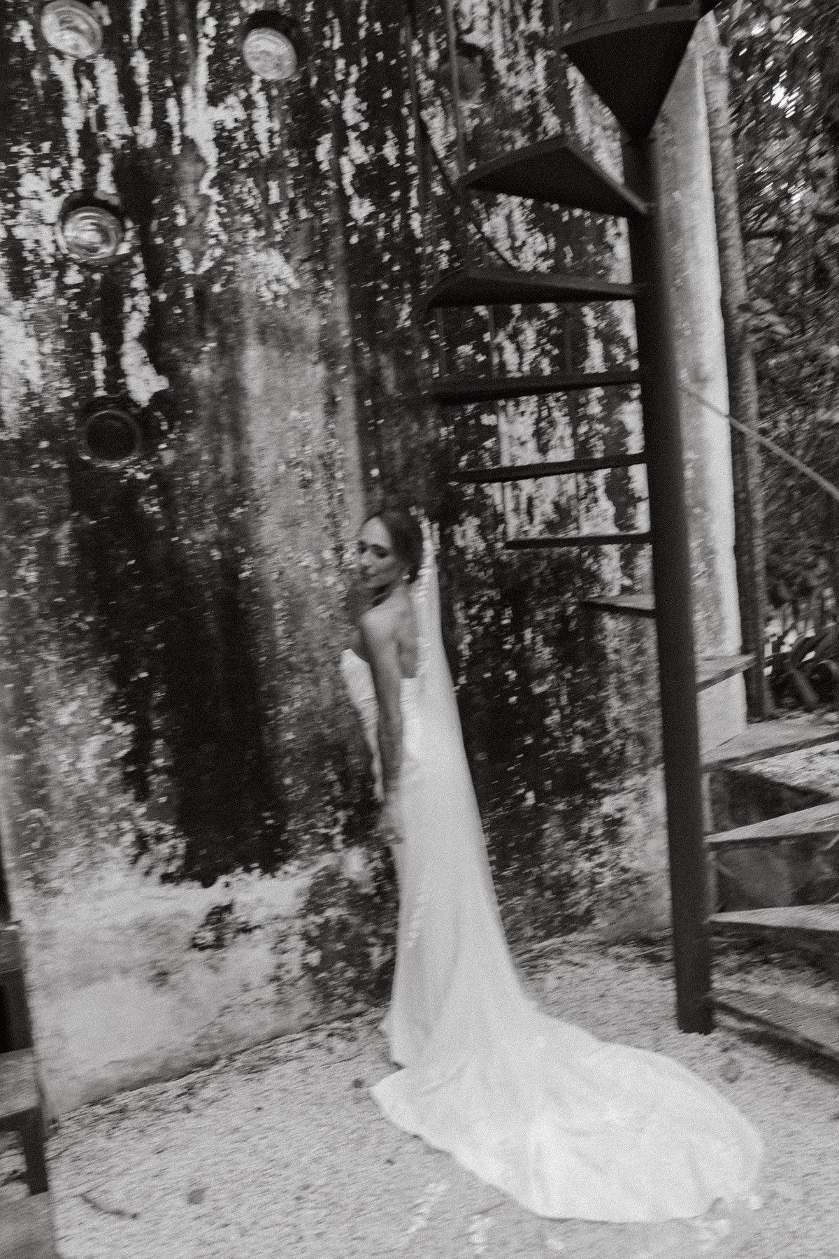 Black and white photo of a bride in a elegant backless wedding gown with long train near rustic stone wall.