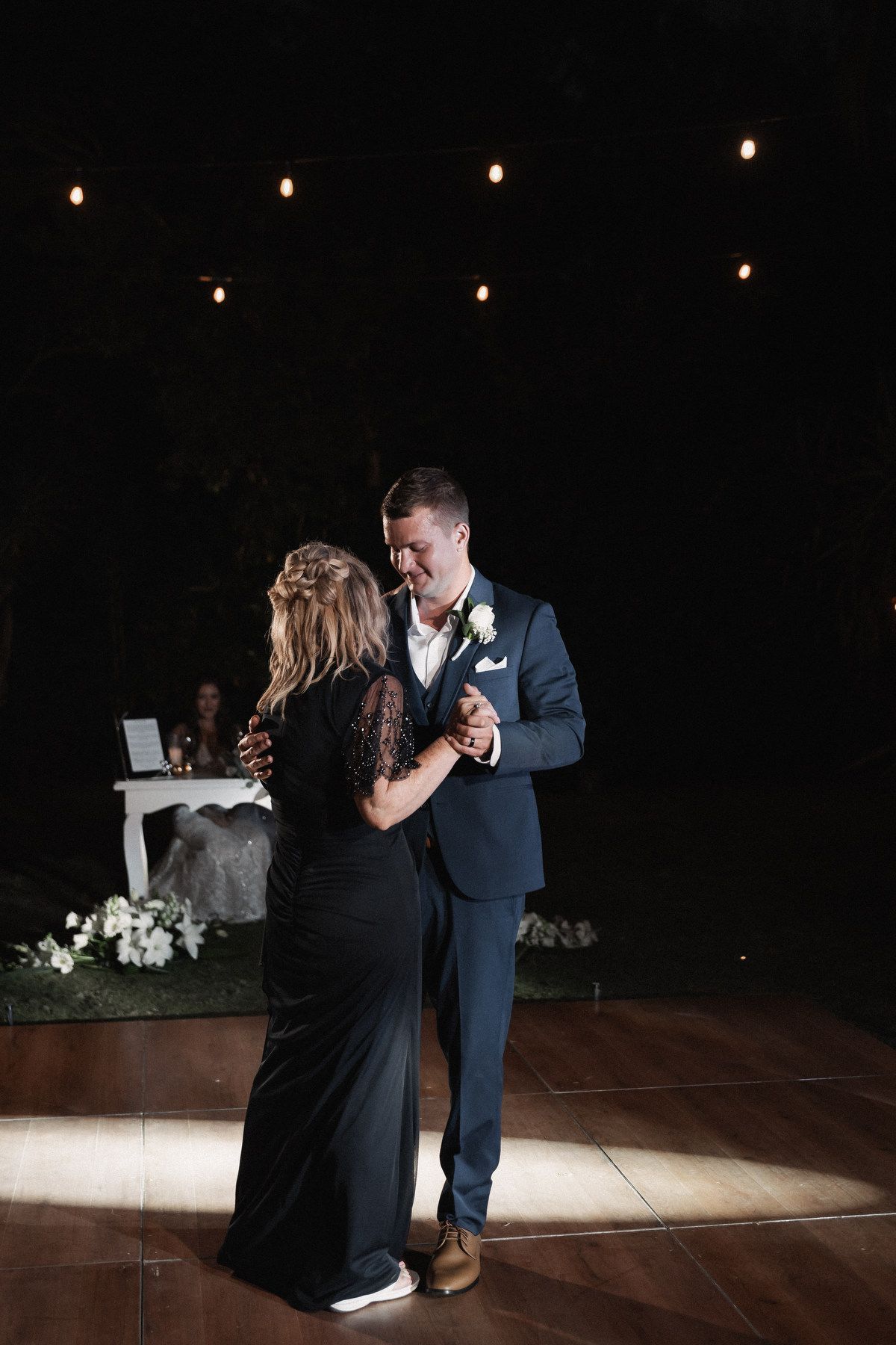 Groom in navy suit shares mother-son dance with woman in black gown under string lights at elegant wedding reception.