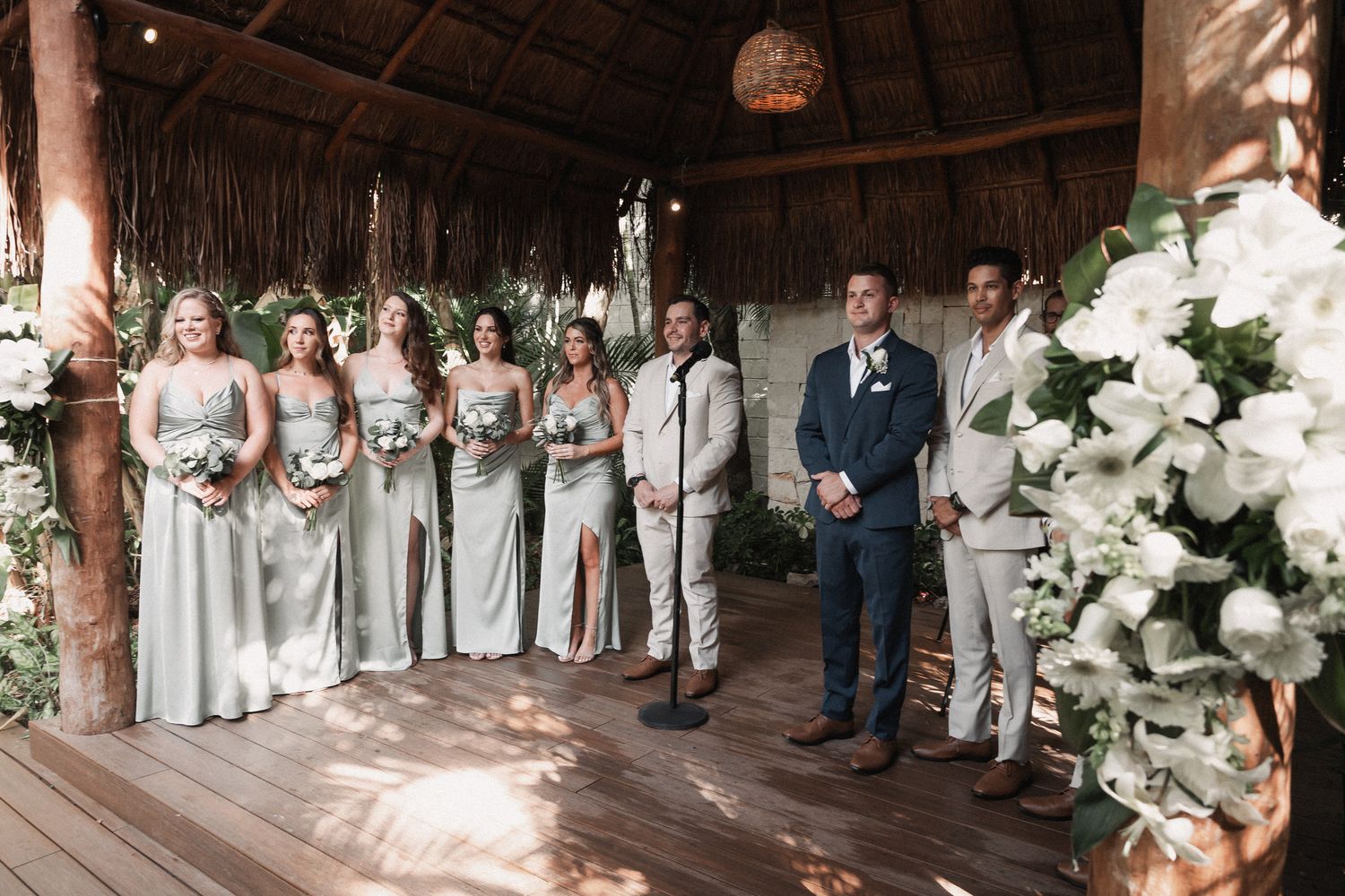 Wedding party stands at tropical palapa altar with sage bridesmaids and tan groomsmen surrounded by white floral arrangements.