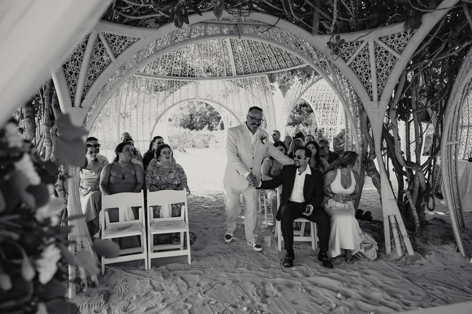 Black and white photo of beach wedding ceremony under decorative gazebo with guests seated.