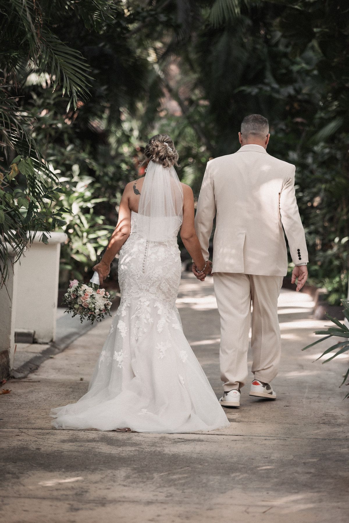 Bride in lace mermaid gown with veil holds bouquet walking hand in hand with groom in beige suit.