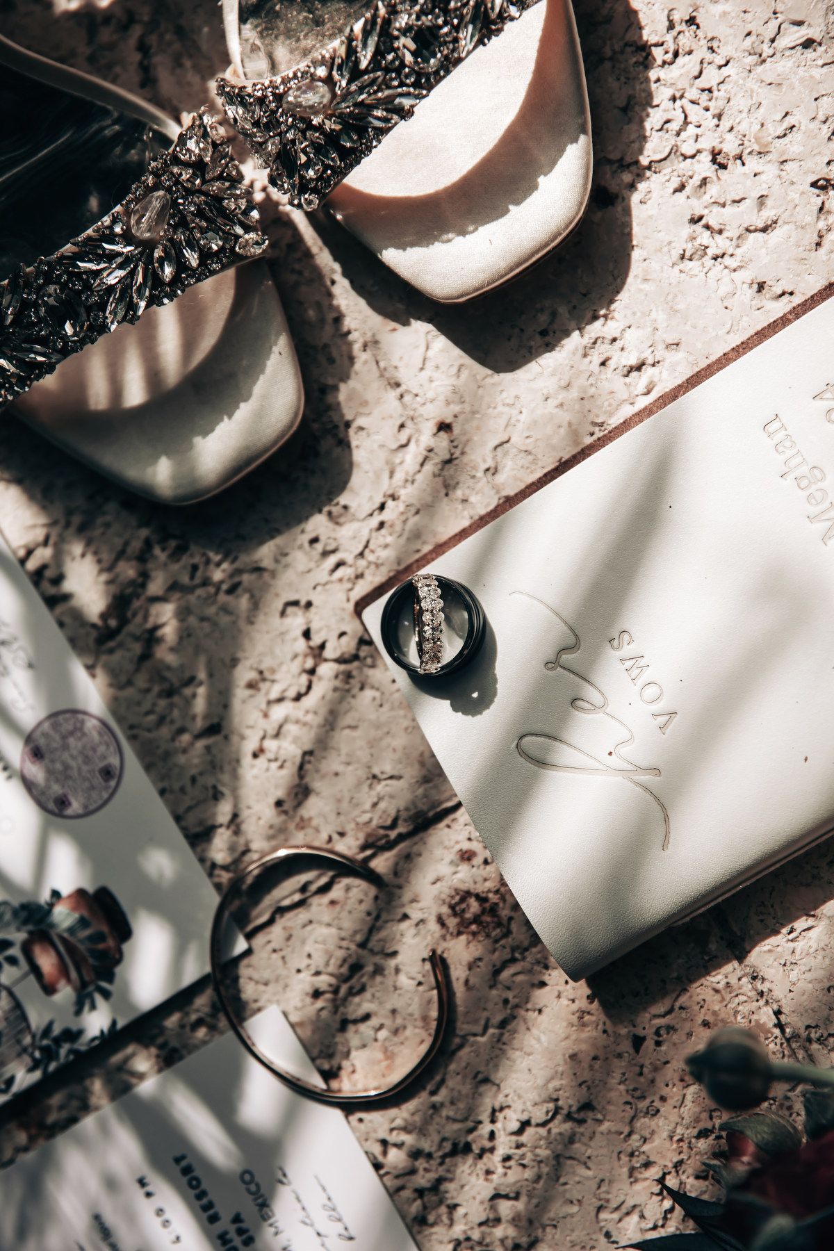 Bridal flat lay with white heels, pearl hair accessories, wedding stationery, and jewelry on textured surface.