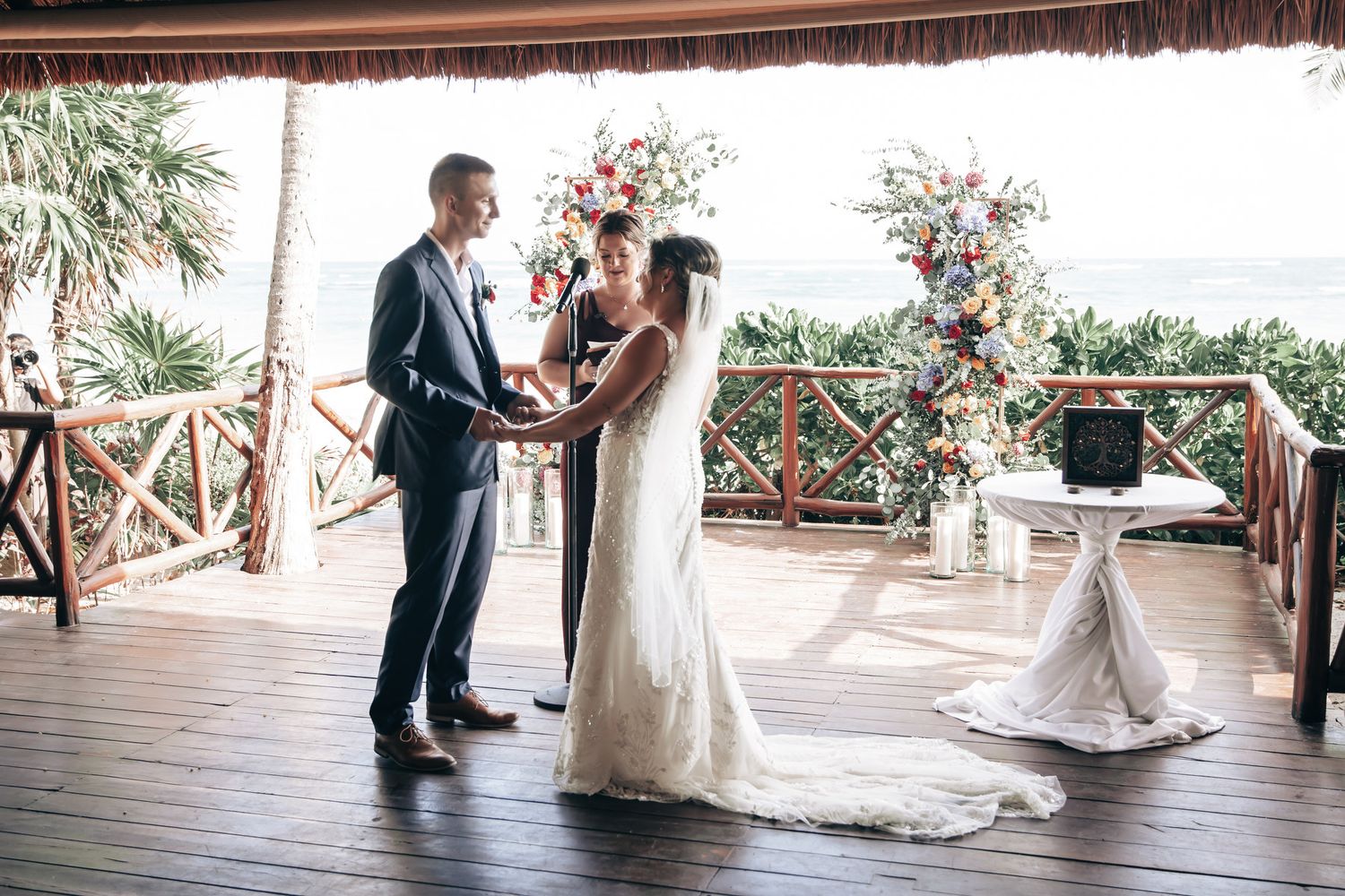 Bride and groom exchange vows at floral altar on wooden deck overlooking tropical ocean scenery.