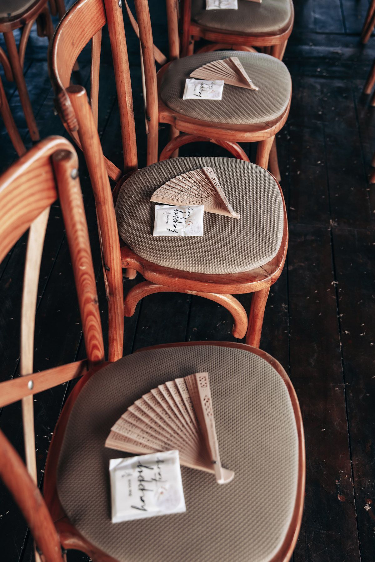 Wooden bistro chairs with cushioned seats displaying paper hand fans and calligraphy cards for a wedding ceremony.