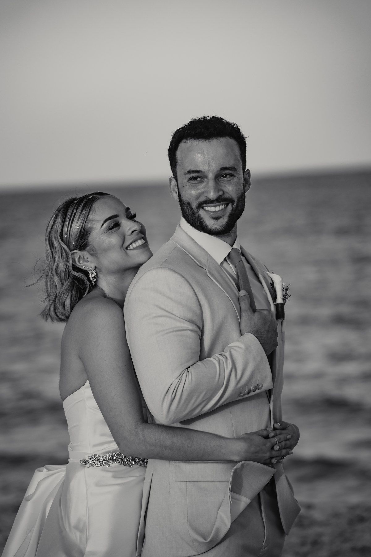 Black and white portrait of bride and groom embracing on beach at sunset.