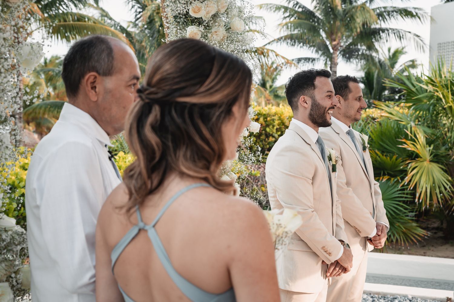 Groom in beige suit smiling at the altar surrounded by groomsmen and bridesmaids at outdoor ceremony.