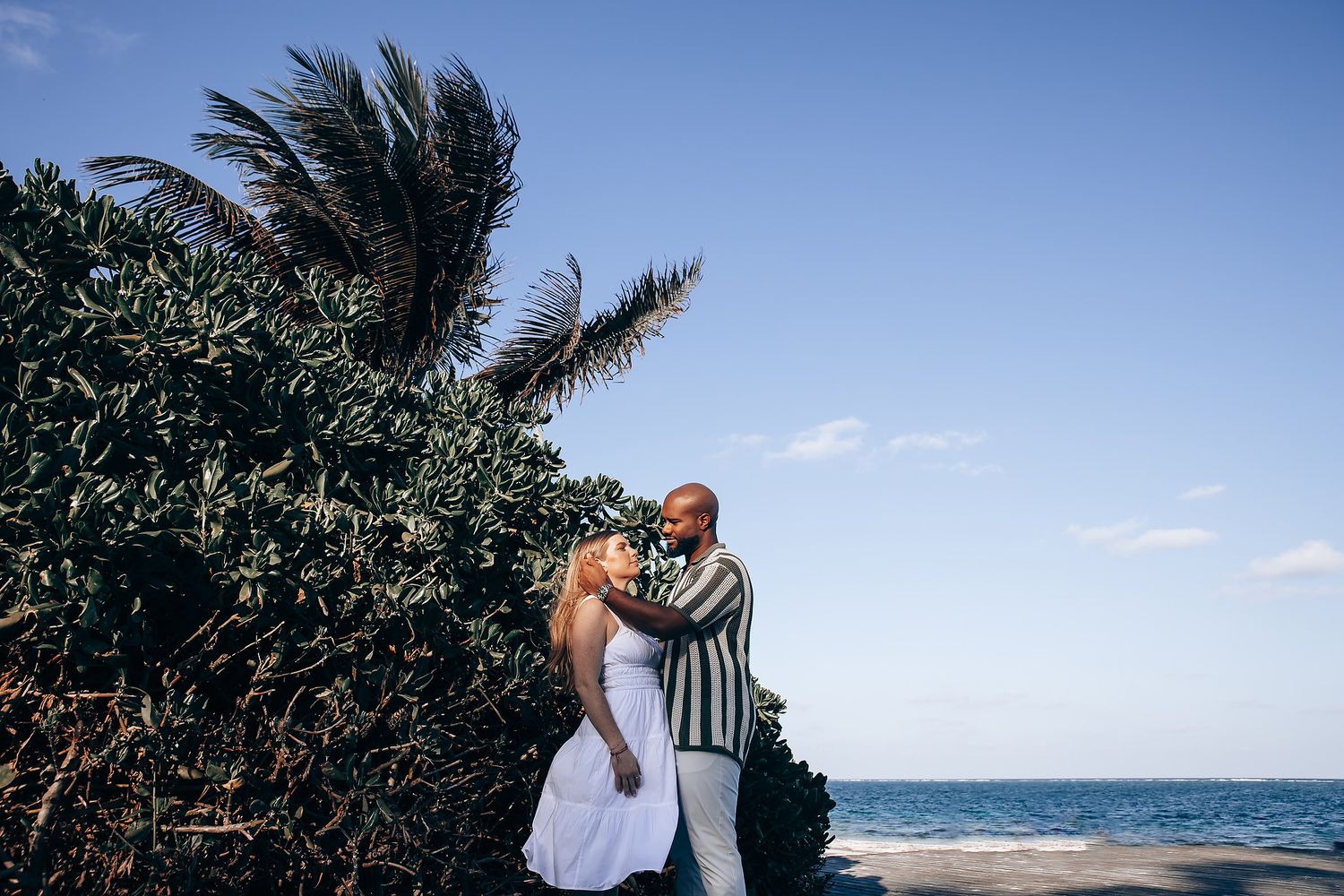 Interracial couple embracing near lush tropical greenery with blue ocean and sky backdrop.
