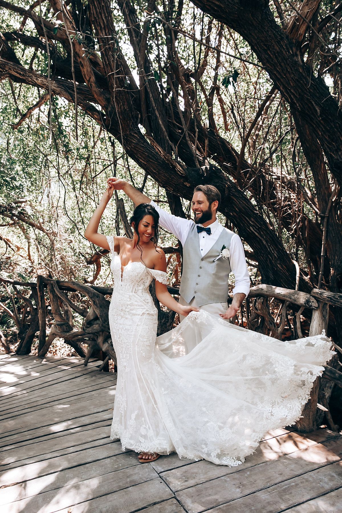 A bride in a flowing white dress twirls on a wooden boardwalk beneath overhanging trees.