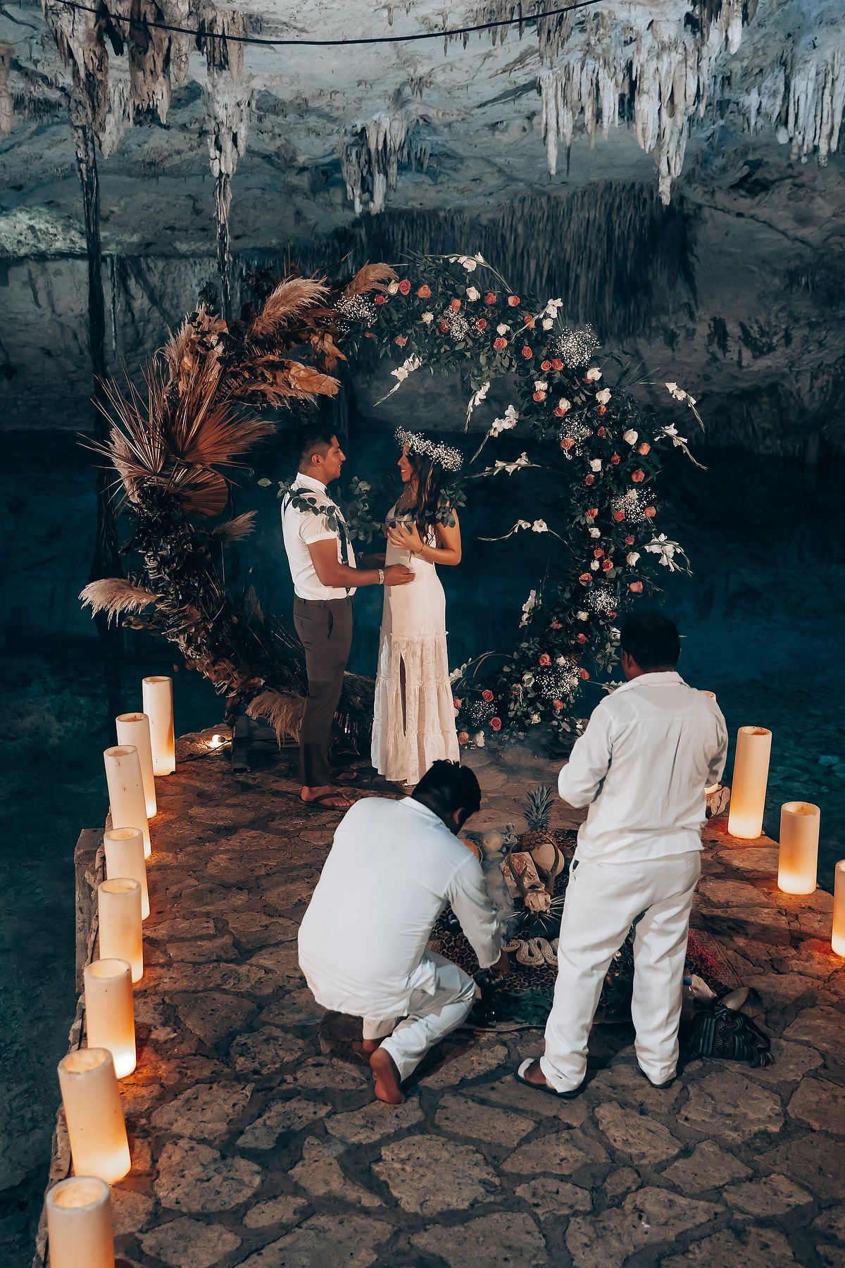 A romantic wedding ceremony setup in a cave with a floral arch and glowing candles along the stone floor.
