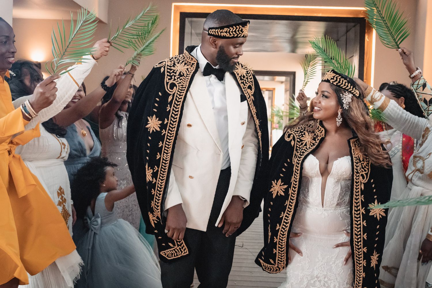 Couple in ornate gold and black traditional wedding attire walking through palm leaf arch with guests.