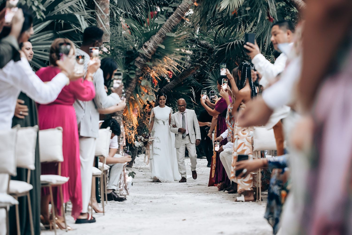 Wedding ceremony aisle decorated with tropical foliage and guests seated on white chairs.