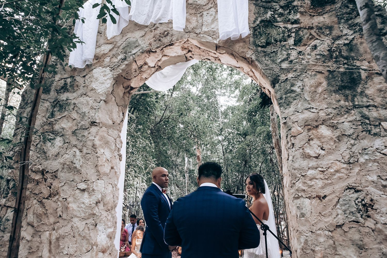 Wedding ceremony takes place in front of a rustic stone wall surrounded by lush greenery and natural light.