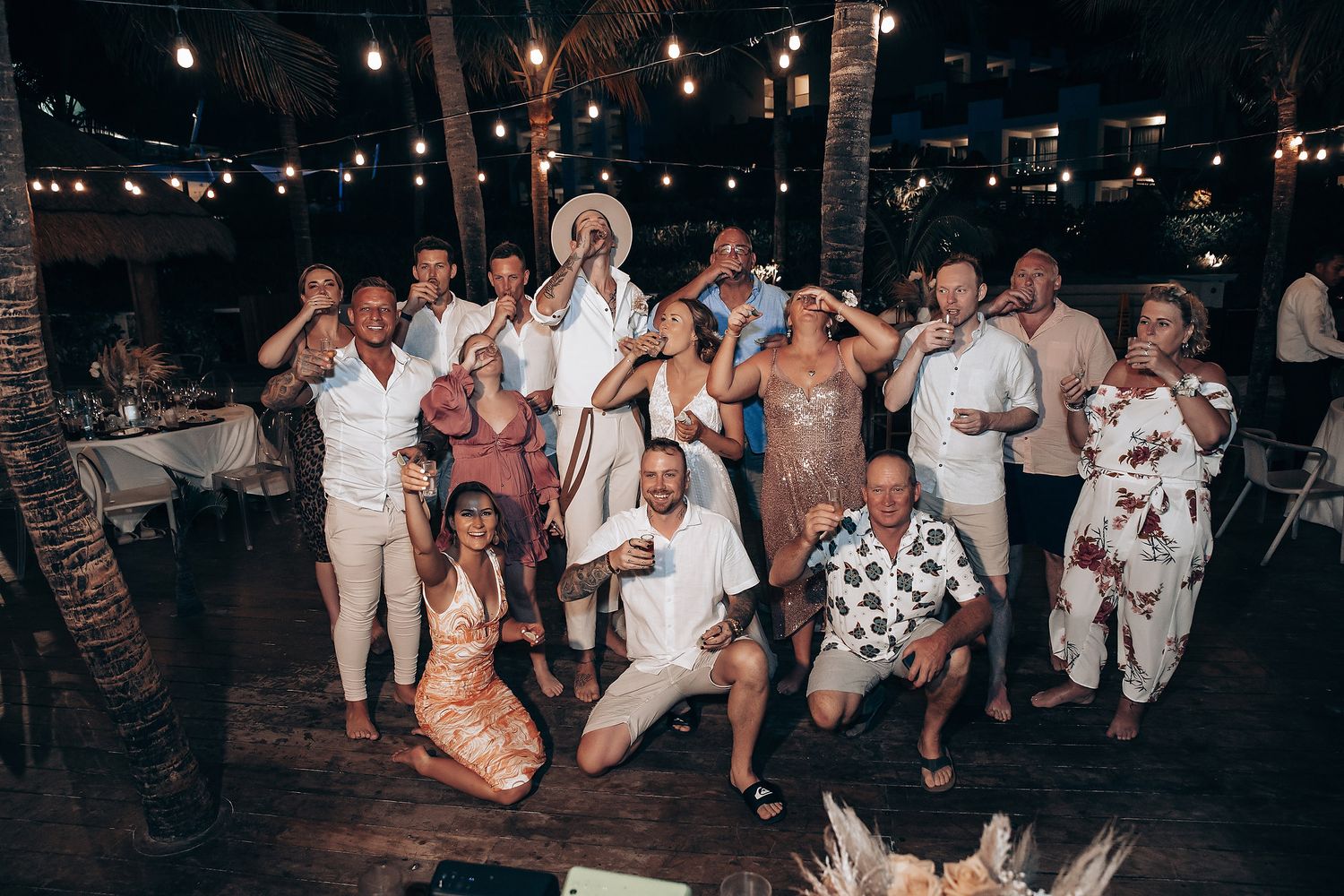 Lively evening reception party with guests dancing under string lights and palm trees.