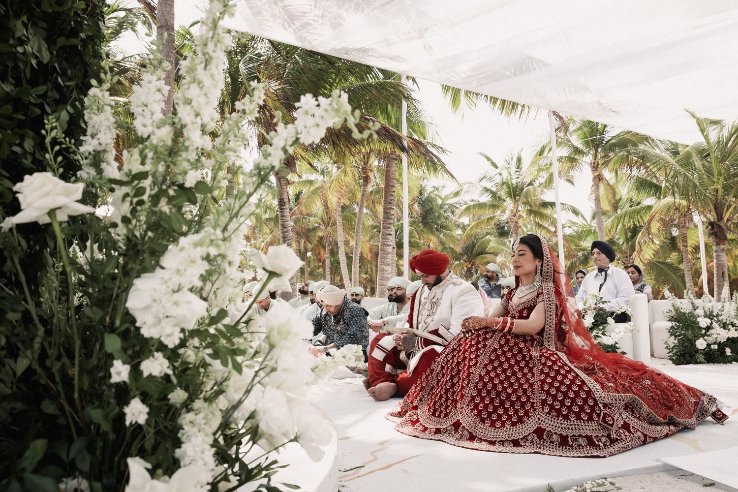 Bride and groom in red attire sit together during outdoor Sikh wedding ceremony with white florals.