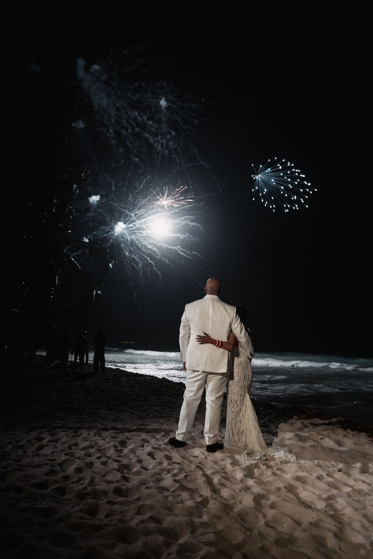 Couple in white attire embracing on beach at night watching colorful fireworks display over ocean.