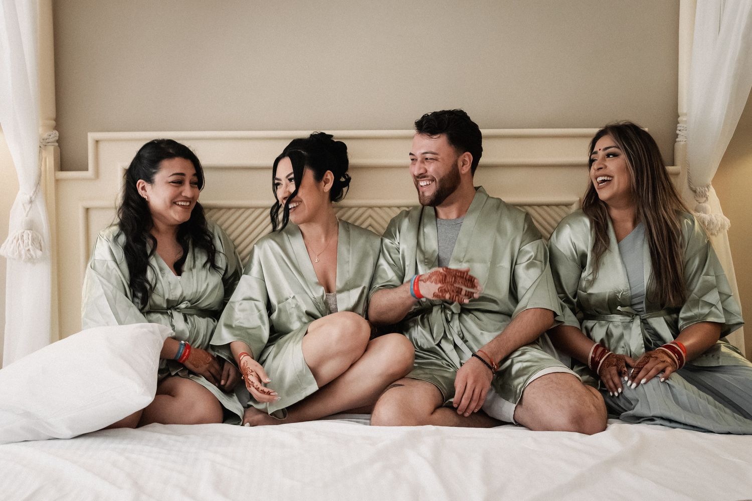 Four friends in matching sage green satin robes laughing together on a bed during bridal party preparations.