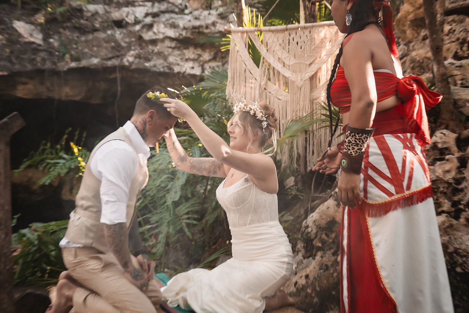 Couple exchanging vows in a tropical cave wedding ceremony with a macrame backdrop and officiant in red dress.