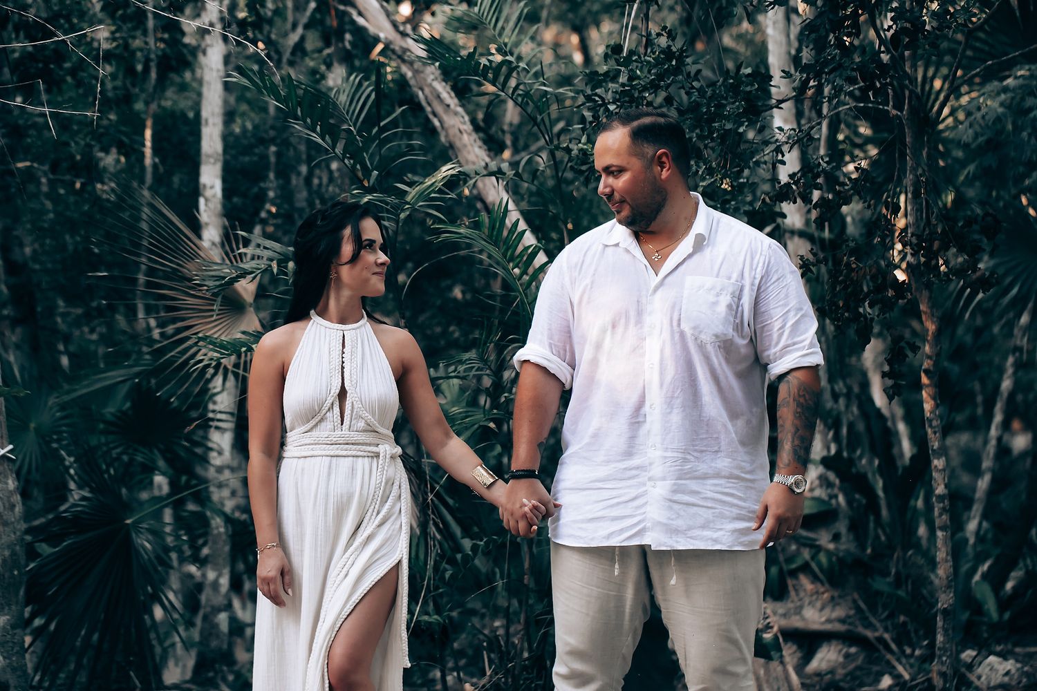 Couple in white attire walking hand in hand through tropical forest setting.