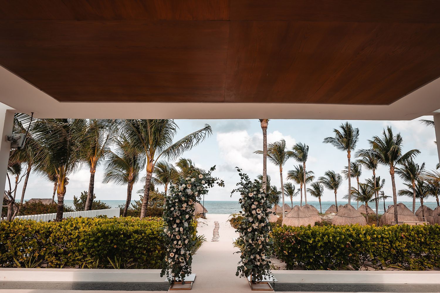 Elegant beachside wedding ceremony setup with floral arrangements framing a path to the ocean, surrounded by palm trees.