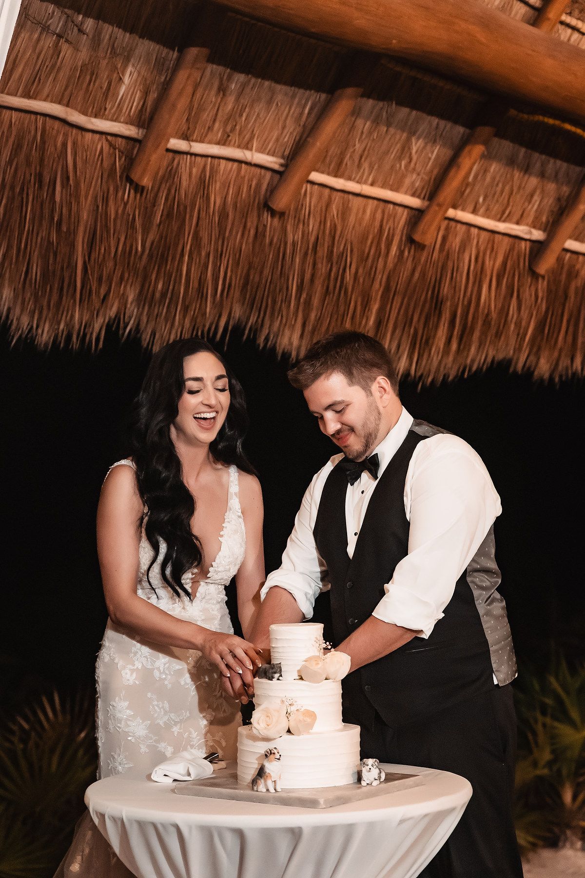 Bride and groom laughing while cutting their white tiered wedding cake under a thatched palapa.