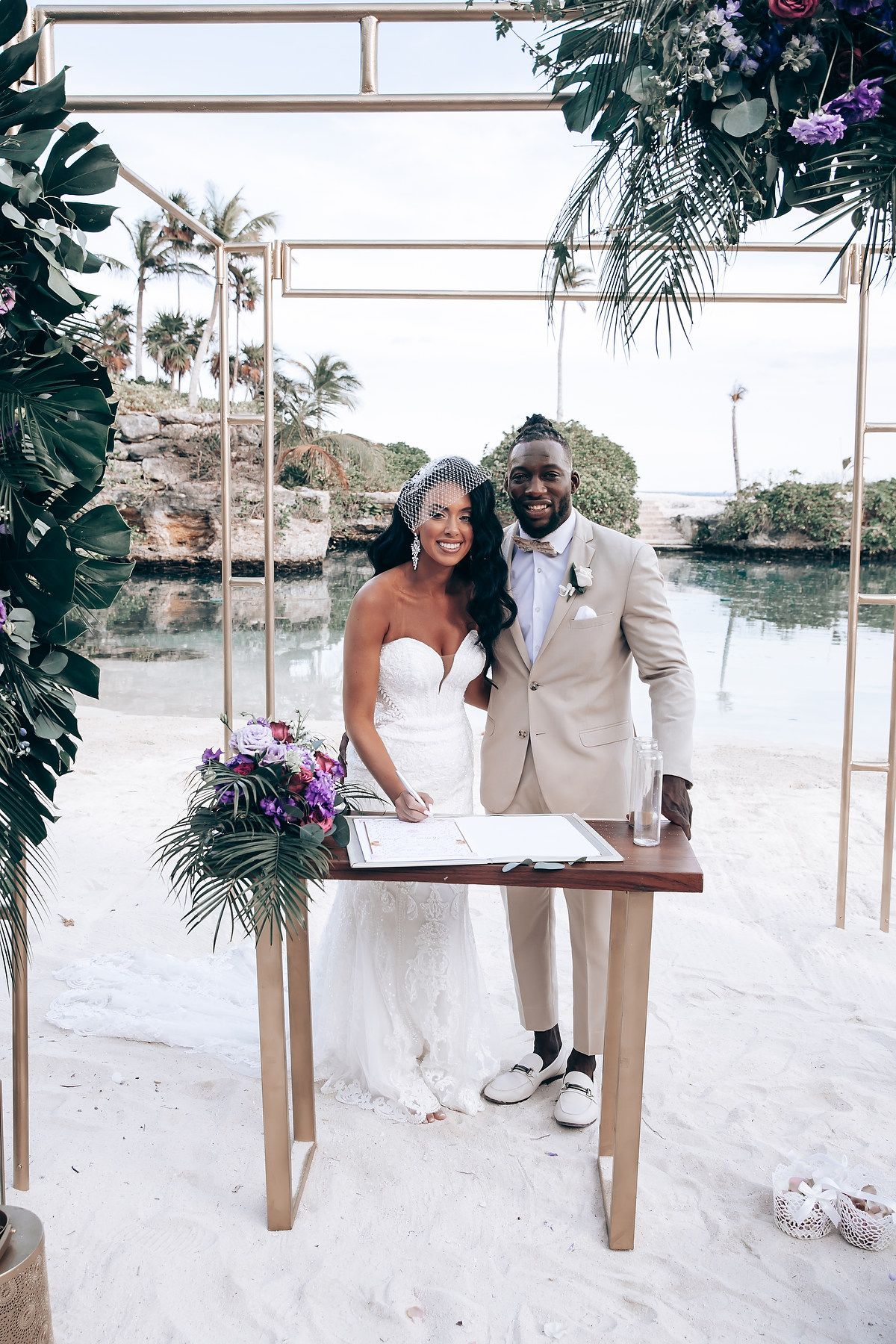 Couple posing on wooden swing frame decorated with tropical leaves on beach.
