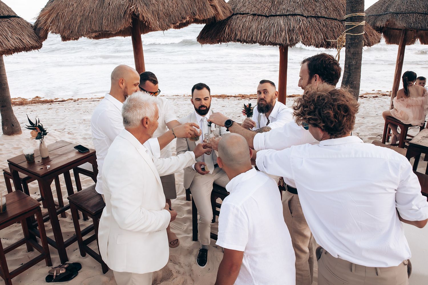 Wedding guests in white attire gather for drinks under thatched umbrellas at a beach reception.