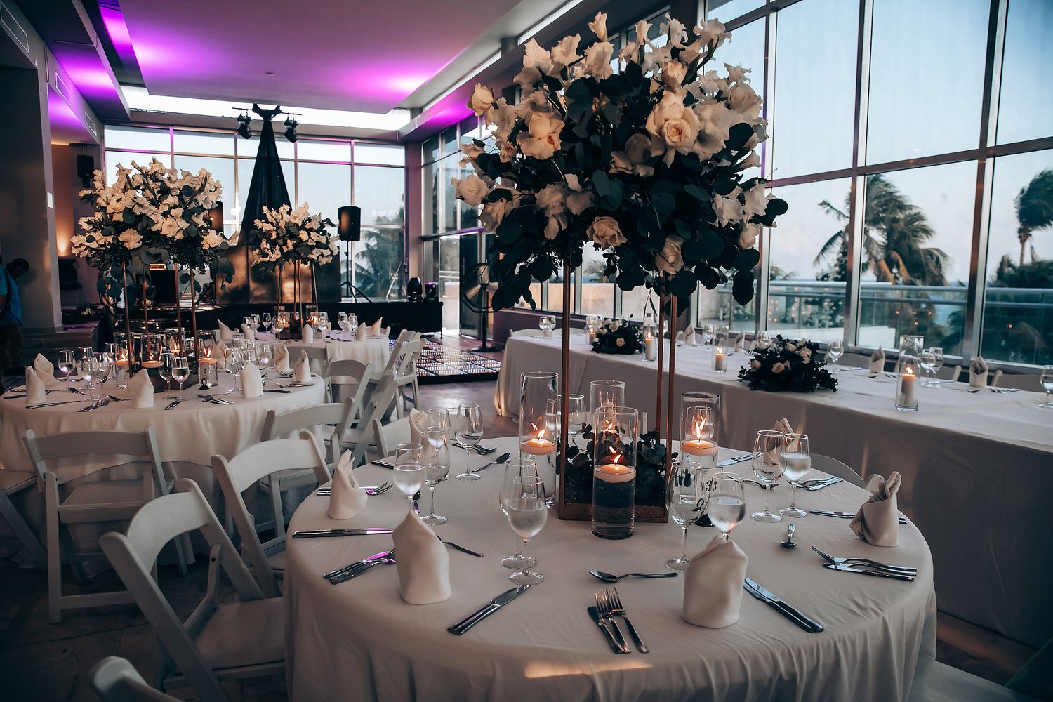 Modern reception venue with elegant table settings, purple lighting, and floor-to-ceiling windows.