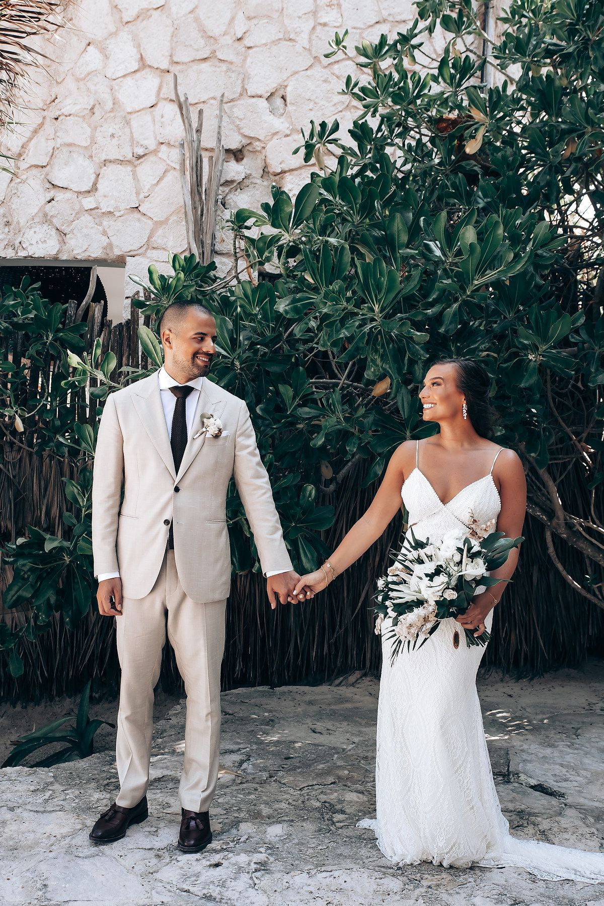 A couple in wedding attire poses against a stone wall backdrop surrounded by tropical foliage.