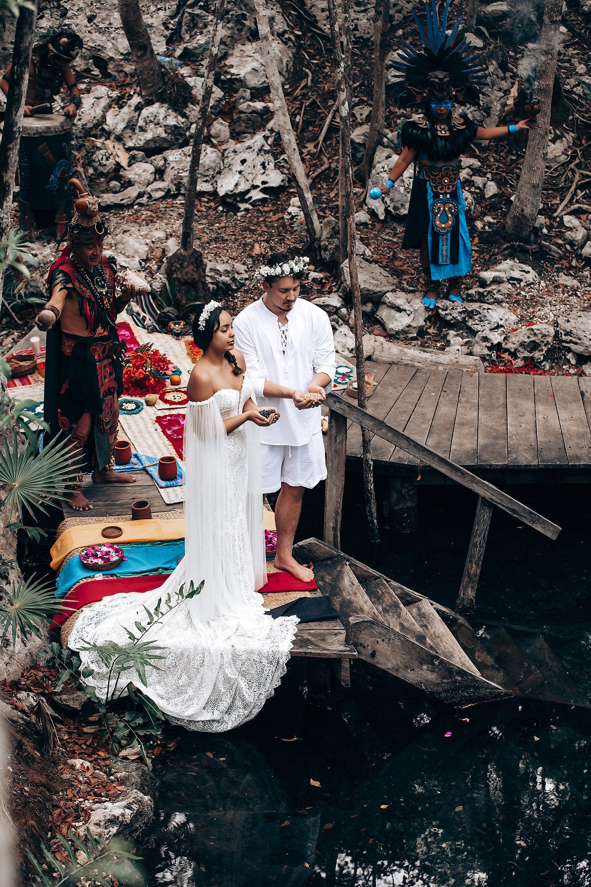 A wedding couple poses on a rustic wooden deck surrounded by lush tropical foliage and dark rocky walls.