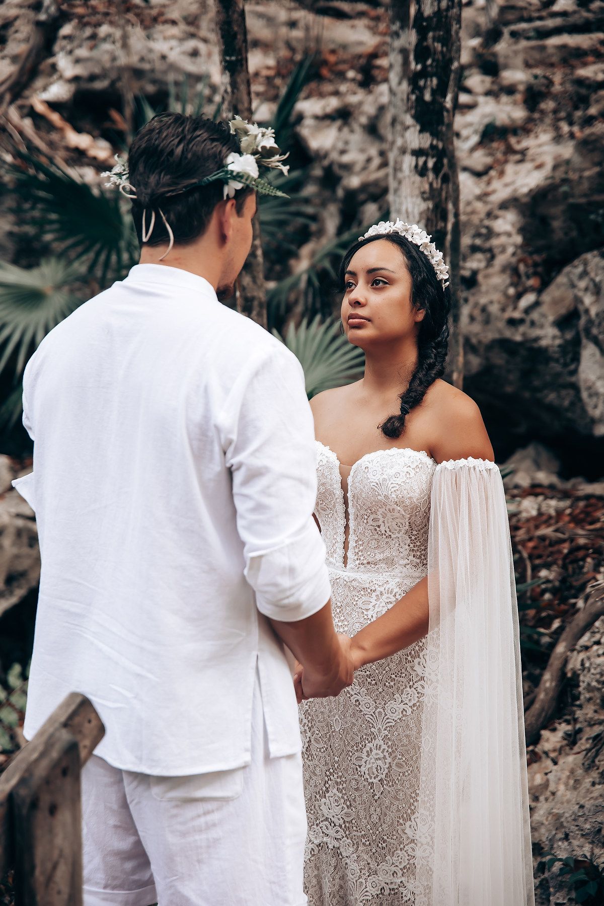 Couple in white wedding attire shares a tender moment on a wooden deck with rocky cliffs in the background.