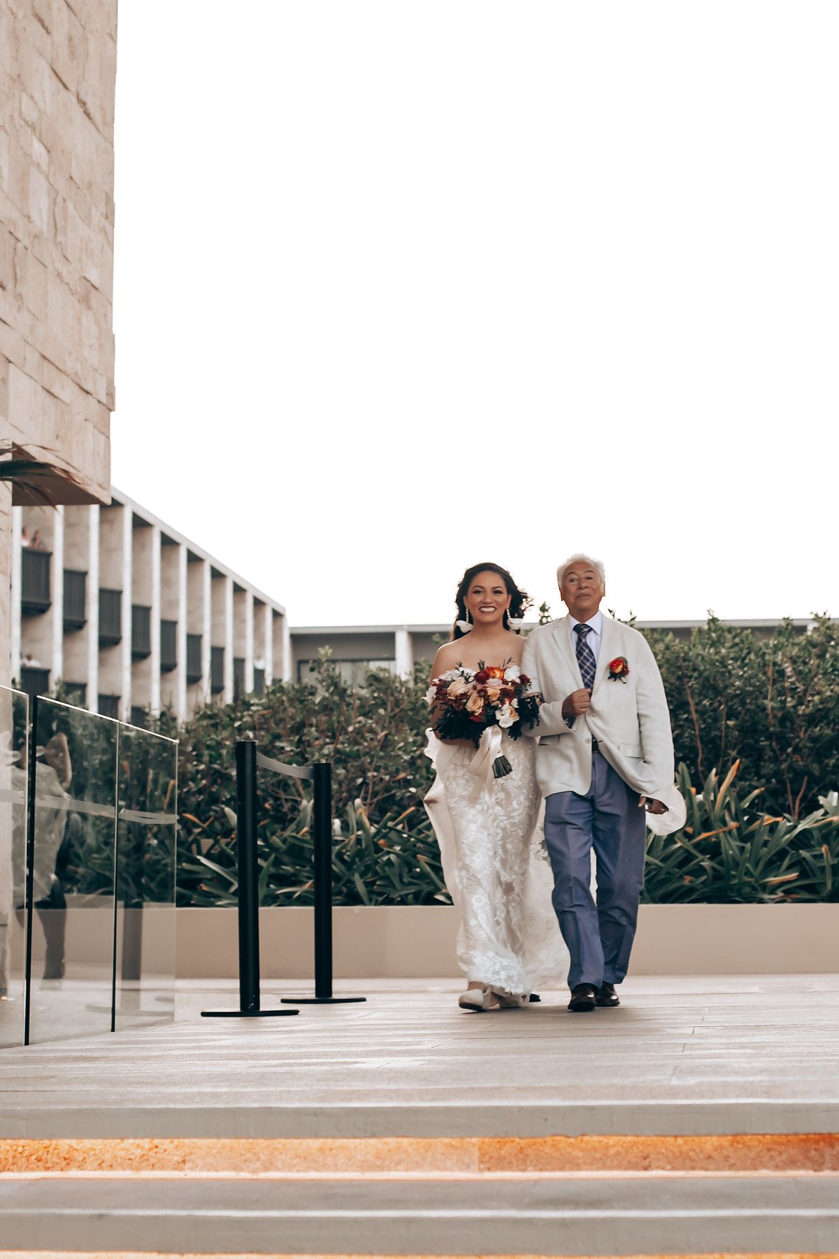 A couple walks together at their wedding ceremony on an outdoor terrace with greenery in the background.