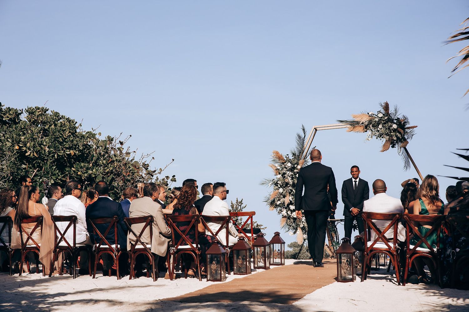 Beach wedding ceremony setup with wooden chairs and tropical decor against ocean backdrop.