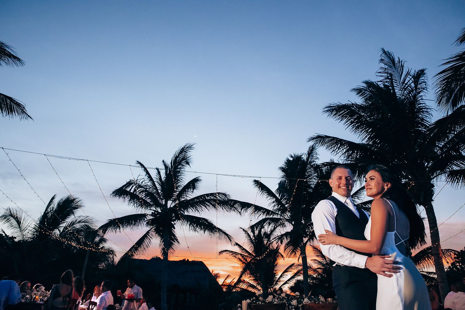 A couple shares a romantic dance at sunset with silhouetted palm trees framing the twilight sky.