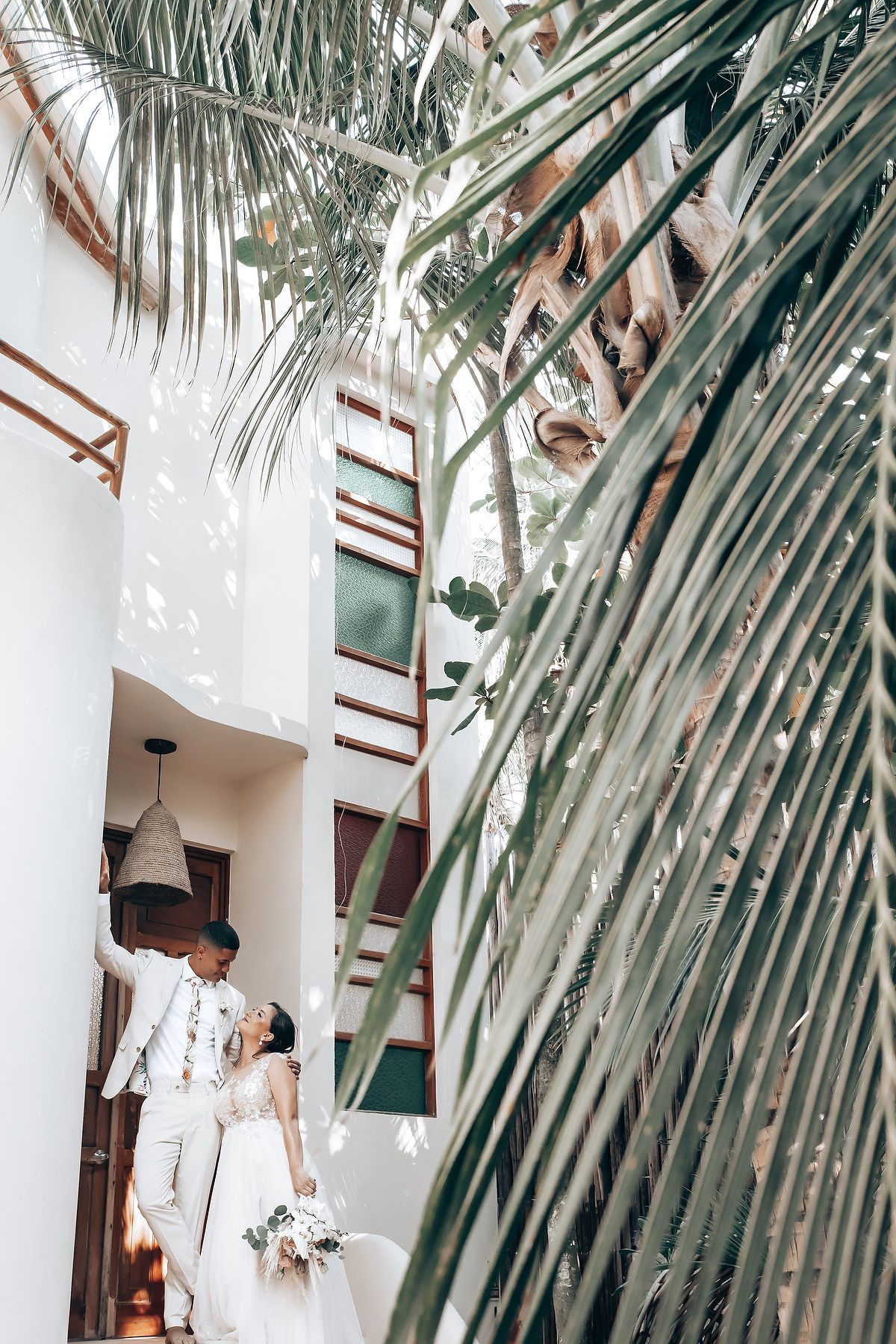 A tropical wedding scene with palm fronds framing a couple in white attire.