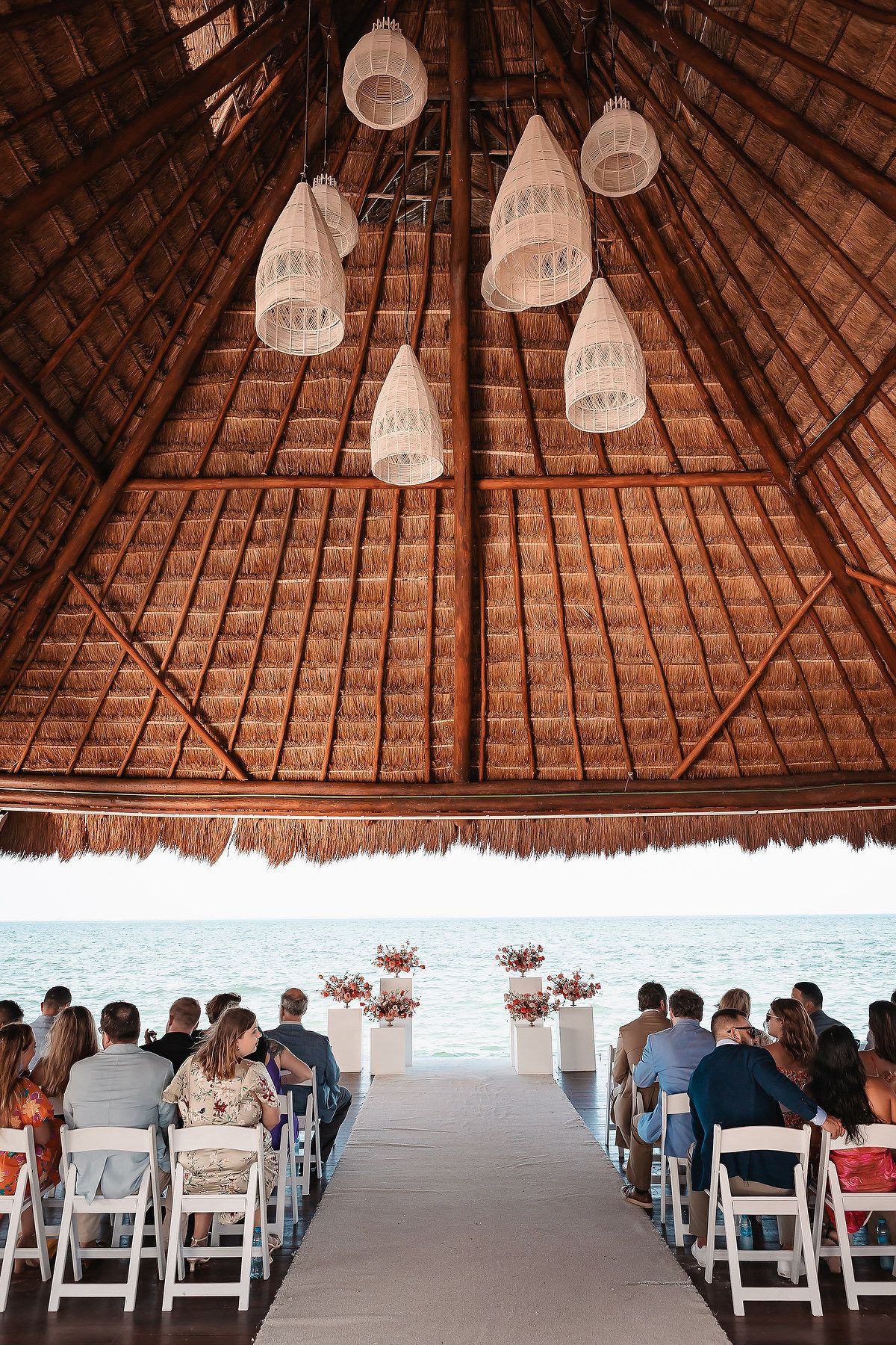 Wedding ceremony setup under a thatched palapa roof with white chairs and ocean view.